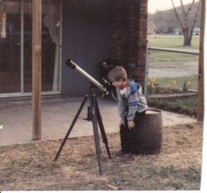 Young boy sitting on wooden barrel peering through telescope.
